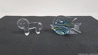 Clear glass lion and colorful blown glass angel fish figurines photographed from front on black textured surface.