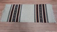 Full view of hand woven rectangular rug with striped pattern and fringed edges on hardwood floor.