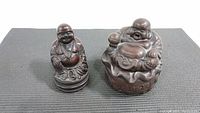 Two metal Buddha statuettes photographed on a black textured mat, showing top-front angled views of the Burner Buddha on the right and the smaller Happy Buddha on the left.