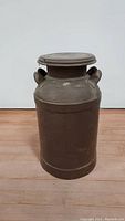 Front and side view of the vintage metal milk can showing the cylindrical body, handles, and lid.