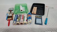 Overview of assortment of paint supplies including brushes, trays, rollers, masking film, and tape arranged on white surface.