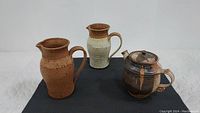 Front view of all three stoneware pitchers, showing shapes, colors, and one lid.