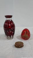Three red glass decorative pieces including a cut glass vase, a red glass egg with gold design, and a small round trinket box