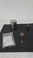Three silver serving ware items placed on a dark mat: container with lid, scalloped spoon, small shaker