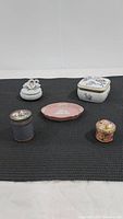 Top view of 5 trinket boxes on black mat showing all items including porcelain doves box, square cherry blossom box, pink Wedgwood-style dish, and two small metal boxes with floral decoration.