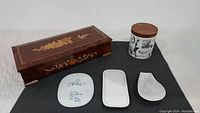 Photo showing all items laid out on a dark cloth, including wooden Thorens music box with floral inlay, porcelain humidor jar with wooden lid, and three small white dishes.
