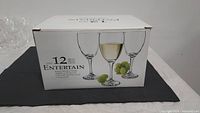 Closed original white box labeled 'Entertain' with image showing 12 wine glasses, box placed on dark mat with some loose glasses visible in background.