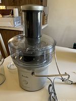 Front view of Breville Juice Fountain juicer showing silver base, clear plastic juice container with black feeding chute and locking arm, power cord visible.