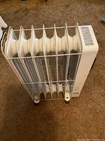Front view of the white oil-filled radiator heater with 7 fins and protective metal grille, standing on brown carpet.