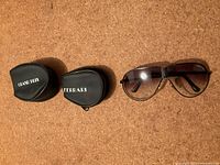 Two folding aviator sunglasses and two black leather-like protective folding cases, one branded Ferrari, the other Grand Prix.