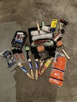 Top-down photo showing full set of painting supplies arranged on concrete floor: roller trays, multiple rollers in plastic wrapping, brushes, gloves in orange packaging, and blue tubes.