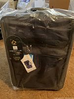 Full view of the black Samsonite luggage wrapped in plastic with original tags attached, showing front zip pocket and handle.