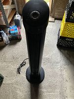 Black Seville upright tower fan standing on floor with power cord visible, surrounded by some boxes and crates.