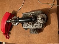 Metal ornate rotary dial phone with black handset and detailed engraving on metal body, photographed on wooden surface.