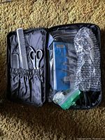 Open zipper case showing grooming tools including black comb, scissors, clipper blades and accessories packed in plastic and bubble wrap.