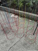 Nine medium sized metal tomato trellises of various colors arranged on concrete ground, shown from above at an angle.