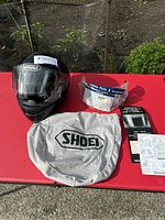 Photo showing black Shoei helmet with clear visor installed, replacement visor in packaging, grey Shoei storage bag on a red surface outdoors.