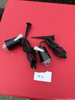 Two black Gemmy Fire & Ice display lights side-by-side on red background. Each light has attached power cord and pointed ground peg stake. Tags and plugs visible.