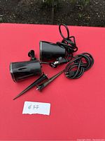 Two black plastic Halloween light projectors laid on a red surface, showing the projector bodies, stakes for ground insertion, and attached power cords with switches.