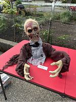 Ghoul Halloween animatronic displayed on a red folding table outdoors, showcasing the full upper body including head, arms, and torso with tattered clothing and skeletal hands.