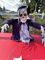Photo showing skeleton animatronic with purple jacket, black hat with purple decoration, skeletal hands outstretched, on red surface with backyard background.