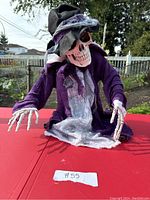 Front view of the Halloween skeleton animatronic seated with outstretched arms. Shows detailed skull with black and gray ragged hat, purple tattered cloak and gray under fabric.
