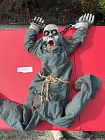 Full view of the Halloween ghoul animatronic laid flat showing shackled hands, frayed clothes, light-up eyes, and rope belt.