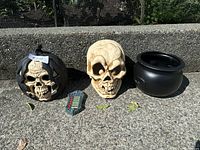 Photo of 4 Halloween props: a black pumpkin skull prop, a large bone-colored skull prop, a black cauldron, and a battery-operated doorbell with buttons, all placed outdoors on concrete.