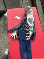 Full body view of the animatronic ghoul dressed in tattered clothing lying on a red surface, showing overall outfit and hands.