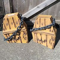 Photo of two wooden coffin-shaped Halloween lawn ornaments with black plastic chain wrapped around each, placed outdoors on concrete against a wooden fence.
