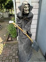 Front view of The Caretaker Halloween animatronic figure standing outside. Shows detailed face, hooded cloak, and shovel held in both hands.