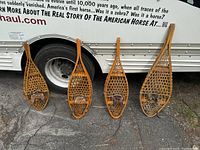 Four pairs of old wooden snowshoes lined up on pavement, showing varying sizes.