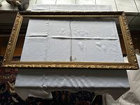Full view of the antique rectangular gesso picture frame lying on a folded white tablecloth, showing its overall size and decorative edges.