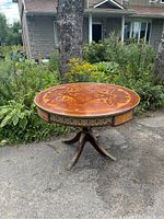 Full view of round antique dining table showing wood inlay and gold-tone trim surrounded by outdoor plants.