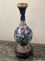 Front angle of the cloisonné vase with visible mythical bird motif and floral designs on a dark blue background. Metal rim shows wear and patina.