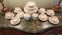 Arranged view of eight place settings including stacked dinner and salad plates, cups on saucers, sugar bowl with lid and creamer on marble surface