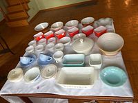 Full view of all Fire King glass items laid out on white cloth on table. Includes bowls, cups, covered casseroles, various sizes and colors.