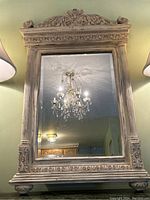 Front view of full floral carved wood mirror showing top leaf and flower design with two lamps reflecting in glass and chandelier reflected.