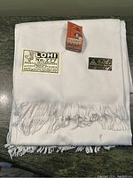 Folded white shawl with fringe, showing manufacturer and product tags including 'LOHI No. 777' and 'Aayushi Creation'.