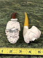 Two meerschaum figural pipes showing detailed carved faces and curved stems beside measuring tape for scale.