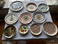 Top view of ten decorative plates laid on white cloth showing varied colors and patterns.