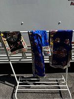 Full view of three kimonos hanging on white rack outdoors under sunlight, showing colorful traditional Chinese embroidery in black, blue, and purple fabrics.