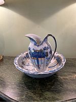 Overall view of the ceramic wash pitcher sitting inside the matching basin on a dark surface under neutral lighting. Shows the design and shape of the pitcher and basin.
