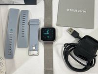 Fitbit Versa smartwatch with metal mesh band, two extra silicone wristbands and USB charging dock with cable beside the original box and manuals.
