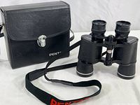 Front view of the binoculars with shoulder strap laying in front of the leather carrying case.