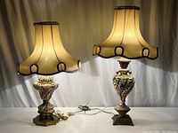 Both vintage china lamps side by side with fabric shades, cords visible, shades beige with dark trim, ornate bases on different metal stands