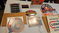 Photo showing various Lionel and MTH decals and signs laid out on a table including large round logos, authorized retailer and service signs, and packets of decals in packaging.