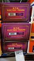 Three stacked purple MTH Electric Trains boxes showing the model names and numbers for Erie Bay Window Caboose, Amtrak Mail Boxcar, and Milwaukee Road flatcar with trailer