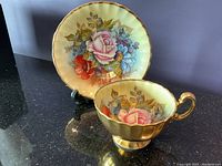 Saucer displayed upright on stand behind teacup showing gold finish and floral motif