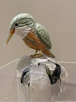 Front left angled view of a carved bird with green, orange, and white stone detail on a quartz crystal base with black mineral inclusions